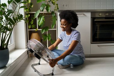 Mutfakta yerde oturan mutlu, genç Afrikalı kadın serinlemek için elektrikli vantilatör kullanıyor. Neşeli siyah kız temiz havanın tadını çıkarıyor. Ev için sağlıklı sıcaklık, sıcak hava dalgaları için termal konfor