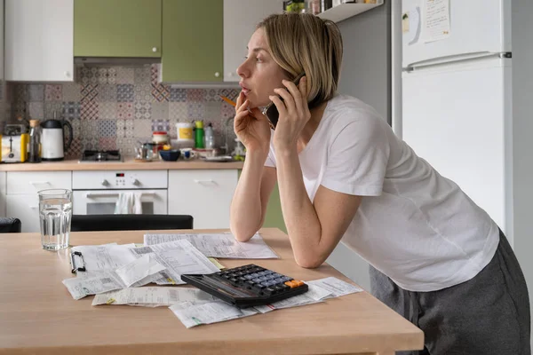 Worried middle-aged European woman housewife managing household finances, making phone call while standing at table with bills and invoices in kitchen. Money management, savings and economy concept