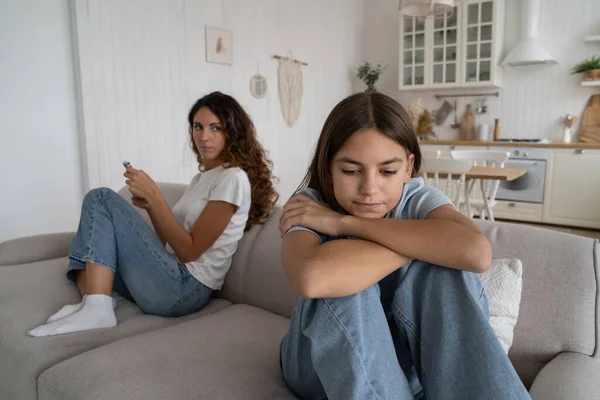 stock image Thoughtful Polish school age girl leans on knees looks into distance feels sad because of house arrest or quarantine. Casual worried Spanish woman sits on sofa behind daughter for teen issues concept