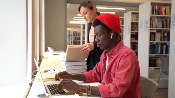 アフリカ系アメリカ人の学生が友人の女性と一緒に図書館で勉強している ヒップスタージョリー男使用ノートパソコン作品のエッセイ 準備のためのテスト試験で女の子学生を探している情報で教科書 — ストック動画