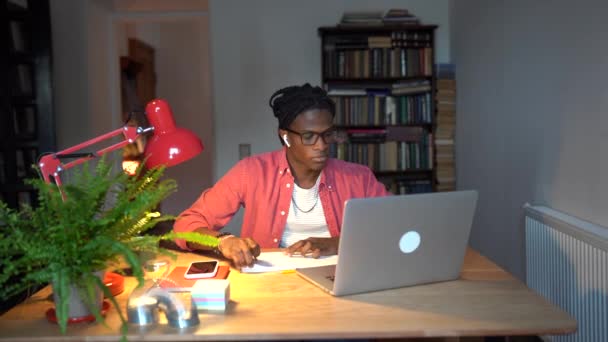 ハンサムなアフリカ系アメリカ人の学生は イヤホンを耳にオーディオタスクは ノートパソコンの画面の情報は 携帯電話に気を取られて笑みを浮かべて読み取りを探しに注意を払う着用 知識を向上させ オンライン学習 — ストック動画