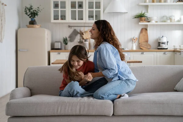 Overjoyed mother enjoying weekend with teen daughter, happy loving mom tickling kid, sitting together on sofa in living room, teenage girl playing with mommy. Fun family activities at home