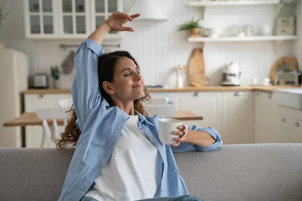 Happy peaceful young woman with cup of drink relaxing on sofa at home, contented female drinking calming tea to chill out, resting on couch with closed eyes and dreaming, enjoying day off alone
