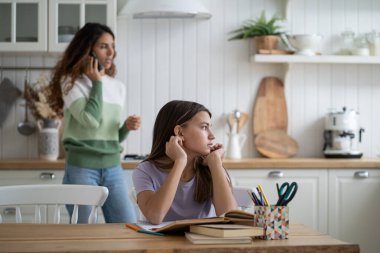 Thoughtful unconcentrated girl look to side doing homework sits at table with textbooks. Woman talking on mobile phone standing in kitchen behind school-age daughter doing extracurricular work clipart