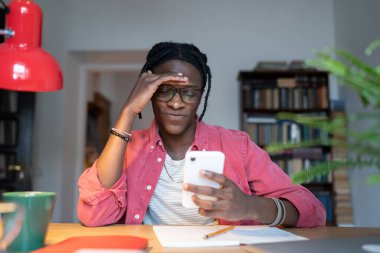 Dissatisfied annoyed African American man sits at table with phone wants to meet girl through internet. Distressed offended Black guy suffers from bullying in comments on photos in social networks  clipart