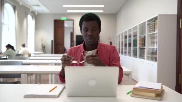 疲れたアフリカの男がノートパソコンとテーブルのライブラリに座って眼鏡を取る目の痛みを感じるストレッチやラバヤシの木 コンピュータと仕事をしながら プロジェクト試験のことを考えている黒人学生 — ストック動画