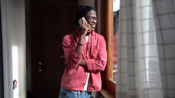 年轻快乐的非洲裔美国人站在窗边 与女友通电话 享受与长距离伴侣通电话的乐趣 微笑的黑人男人在家里用手机和朋友聊天 — 图库视频影像