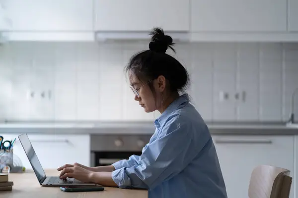 アジアの少女フリーランスの作家で ラップトップでタイプする眼鏡を身に着け 現代のキッチンで机に座り 自宅から遠隔操作しながら集中し 生産性を維持しています フリーランスライティング — ストック写真