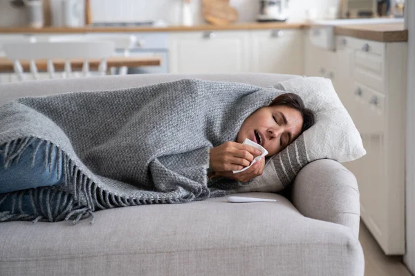 Young woman lying under blanket on sofa coughing and sneezing, using paper tissue to blow her nose. Unhealthy female caught cold or flu, resting on couch, suffering coronavirus. Cold homes and health