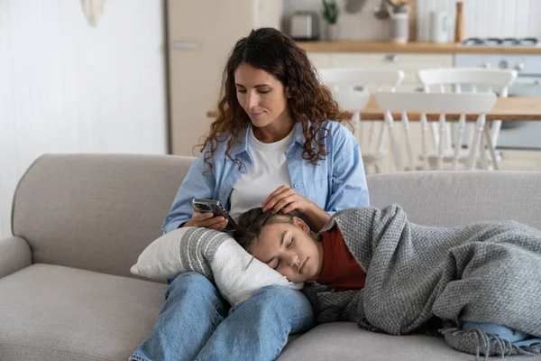 Tired carefree teenage girl sleeping lying on mother lap and wrapped in gray blanket after hard day at school or long walk. Positive woman with mobile phone sits on sofa in home interior with child