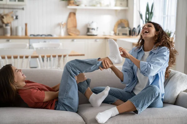 Mother-daughter fun activities at home. Overjoyed mother laughing out of loud while tickling feet of daughter, joyful mommy and teen girl enjoying spending time together, having fun, relaxing on sofa