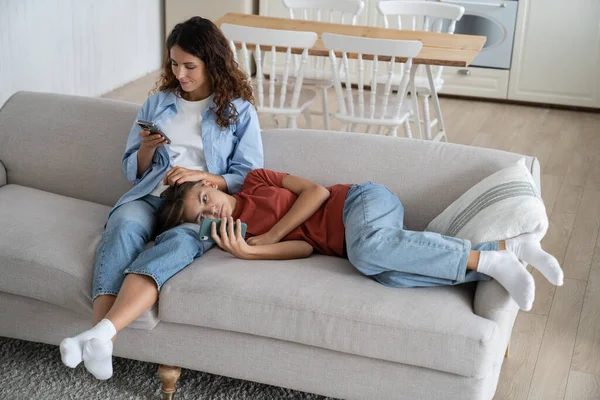 Gadget-addicted family mom and child using smartphones while relaxing together on couch at home, mother and teen daughter resting on sofa using smartphones. Screen time for adults and children