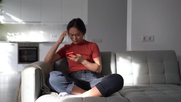 伤心的亚洲女孩一边扔掉手机 一边通过短信与男朋友分手 焦虑的女人在手机上听到坏消息 应付分手 坐在家里的沙发上 — 图库视频影像