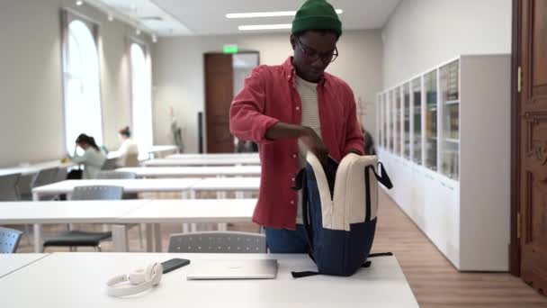 アフリカの若い学生の男は 図書館を出る前に ノートパソコンや教科書をバックパックに入れて勉強を終えた 同僚のスペースで仕事を終える黒人のフリーランサー — ストック動画
