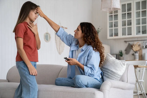 Concerned worried mother checking high body temperature of daughter, touching forehead. Woman mom freelancer holding smartphone listening to child health complains, working from home with sick kid