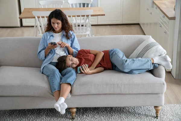 Family mother and teen daughter using mobile devices surfing internet while spending at home, parent and child resting on sofa and using smartphones. Smartphone addiction and family communication