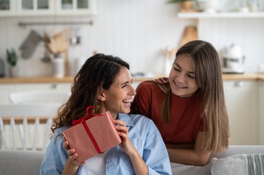 Laughing contented woman babysitter received gift from teenage girl posing near sofa in living room. Cheerful kind daughter gives mom gift on eve of mothers day or on wedding anniversary of parents