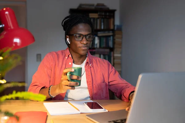 Young African American millennial guy student using laptop for studying and drinking tea, sitting at desk attending online classes in comfortable home environment. Self-education and e-learning