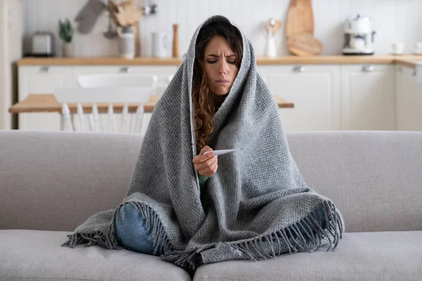 Sick unhappy woman frustrated looking at thermometer having cold during winter walk or work on frost. Diseased sad girl wrapped in blanket has chills and stress sits on sofa in unheated apartment