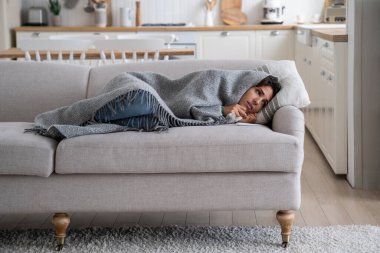 Cold at home. Sick young woman lying under wool blanket on sofa in winter season, staying warm in freezing apartment, ill unhealthy female covered with plaid resting on couch. Central heating problems