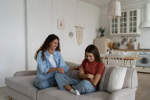 Positive mother and teenage offended daughter sitting on couch at home reconcile after fight, mom making peace with teenager, adolescent child avoid talking with mommy, parent communicating with teen
