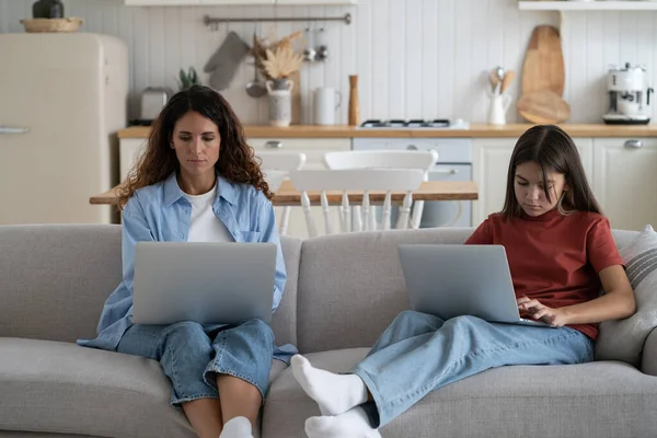 Mother and teen girl child sitting on sofa in living room using laptops, family spending time in front of screen. Mom working remotely while her daughter studying online. Families and technology use