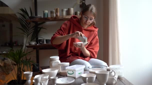 若い女性陶芸家が粘土製品を研磨し 焼成する前に陶器を研磨し 居心地の良いスタジオやワークショップで職場に立っている間 ガラスのための手作りの陶器を準備しました 小規模工芸事業 — ストック動画