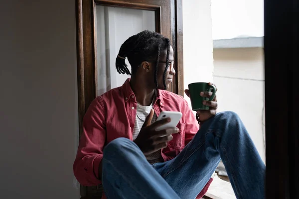 Young smiling African guy holding smartphone and cup of tea enjoying lazy weekend day at home, sitting on windowsill and looking out window. Calm black man turning off phone to spend time in silence