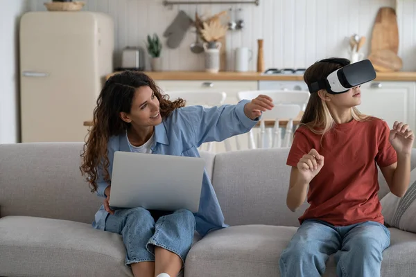 Families and new technologies. Smiling work-at-home mother sitting on sofa with daughter wearing vr helmet exploring virtual reality. Curious child teen girl testing AR technology for education