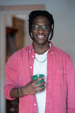 Happy optimistic young African man in glasses holding cup of tea and looking at camera with smile while spending leisure time at home. Joyful black guy freelancer having coffee break. Selective focus