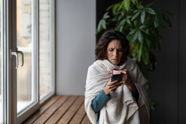 Sick young woman use smartphone consulting with medical doctor online. Unhealthy girl feel unwell suffer from seasonal flu, infection or virus fever and sore throat drink hot tea covered in blanket