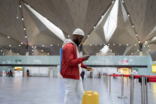 stock image Thoughtful African American traveler man using smartphone at airport terminal, travelling with luggage bag. Passenger guy waiting her flight, checks time of flight online, standing in railway station