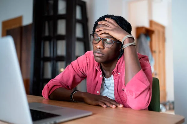 Oppressed depressed African American man closed eyes sits at table with laptop stressed due to lack interest in work. Sad guy in home office feeling burnout and lack of motivation due to low salary