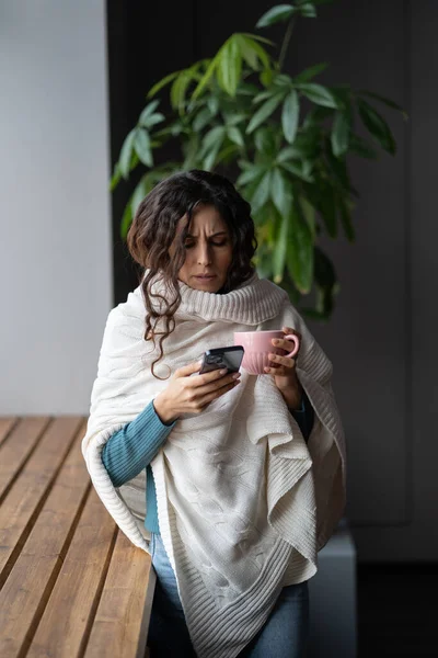 Young unhealthy woman covered in knitted plaid suffering from cold or flu writing sick day email message via smartphone while standing near window at home, sick female letting boss know about illness