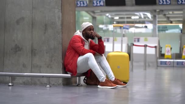 厌倦了的非洲裔美国人在机场心烦意乱 他的航班延误了 焦虑不安的男性旅行者坐在空荡荡的候机楼里 提着行李等着飞机 机场通宵通勤的精疲力尽的家伙 — 图库视频影像