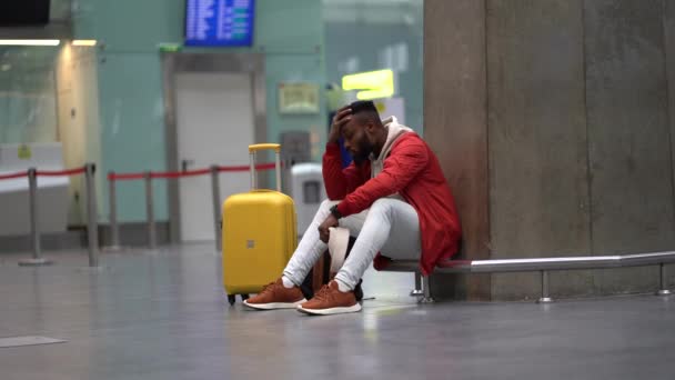アフリカ系アメリカ人の悲しい男が空港で動揺している彼の飛行は遅れている 空のターミナルに荷物を持って座って飛行機を待っている落ち込んでいる旅行者の男性 空港での長い夜の接続の疲れ男 — ストック動画