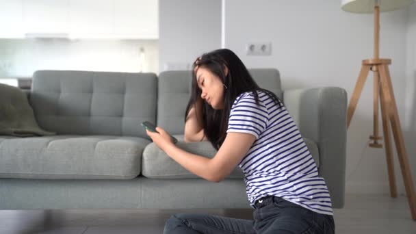 忧心忡忡的亚洲女人坐在沙发旁边的地板上 希望能在电话里看到她的留言 而不是等待男朋友打来的电话 在社交媒体的应用中 不快乐的日本女孩沉溺于喜欢的东西 — 图库视频影像