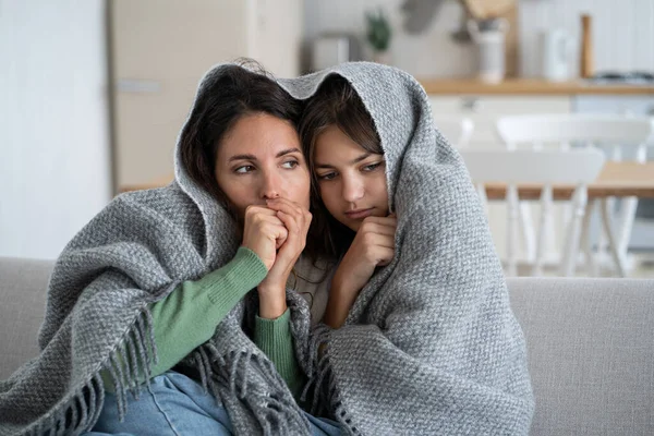 Unhappy frozen woman with daughter are sits on couch cuddling up to each other wrapped in warm blanket. Discouraged depressed family of mother and teen girl suffer due to lack of funds to heat house