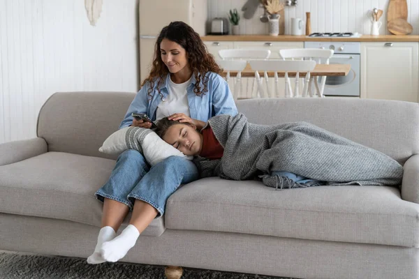 Caring happy mother using phone and stroking teenage girl falling asleep on knees. Cheerful smiling lady in casual clothes is sits on sofa in living room with sleeping daughter wrapped in blanket