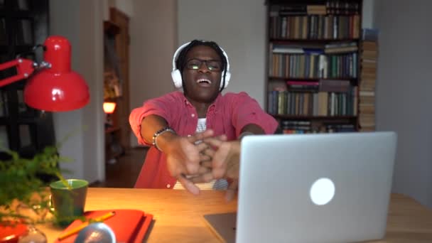 長い仕事の後に暖まる陽気な正のアフリカ人男性は 自宅のオフィスでノートパソコンとテーブルに座っています 楽観的な疲れ黒の男Seoオプティマイザーは マネージャのタスクを完了した後にヘッドフォンを脱いで — ストック動画