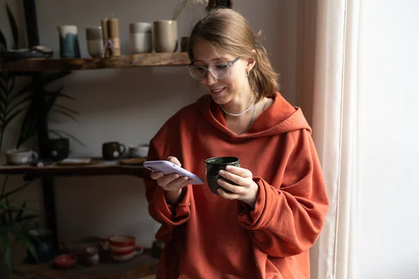 stock image Young happy woman ceramic shop owner using phone and drinking tea, chatting with clients online. Female ceramist taking break while working in cozy pottery studio, potter resting at workplace