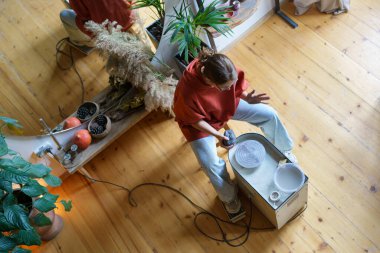 Young woman amateur ceramic enthusiast using electric pottery wheel machine while making ceramics at home, view from above. Female ceramist shaping clay, working in studio. Pottery-making techniques clipart