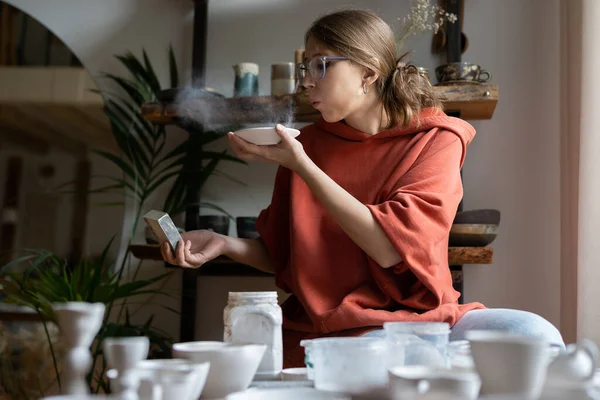 stock image Woman potter working in cozy studio using sandpaper, polishing pottery, female ceramist business owner sanding ceramics, blowing away clay dust while smoothing surface of earthenware