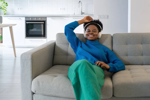 Happy relaxed African woman in headphones closing eyes listening to audio training or podcast of favorite comedian. Positive cheerful black girl relaxing enjoying music sits on sofa in apartment