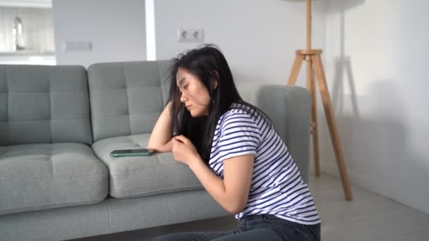 忧心忡忡的亚洲女人看着智能手机屏幕 等待男朋友发短信或打电话 坐在家里的地板上 分手后 伤心的女人在打电话时心情低落 — 图库视频影像