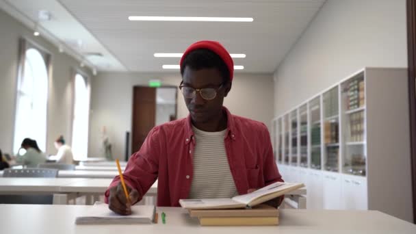 図書館で勉強したり 本を読んだり メモを取る眼鏡をかけている才能のある若い黒人科学者 アフリカ系アメリカ人の男性学生に焦点を当て 机の上に座って研究概要を書く — ストック動画