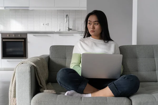 Young thoughtful Asian woman holding laptop on laps looking in distance thinking about personal issues while working from home. Pensive female sitting on couch waiting for important email