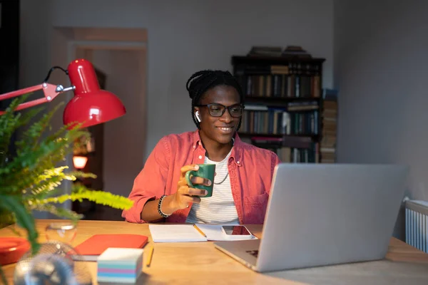 Smiling African student guy in wireless earbuds holding cup of tea coffee watching online lectures at home while studying remotely, learning online for free, looking at laptop screen. Selective focus