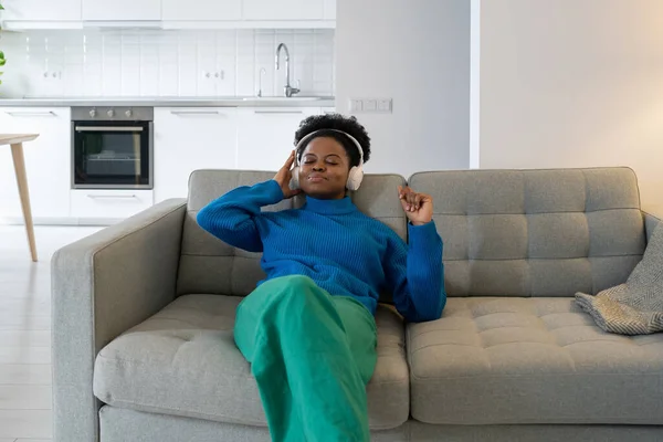 stock image Carefree cheerful African woman listening to music holding headphones to ears during playback playlist. Smiling calm black girl closing eyes resting after working day sits on sofa in living room