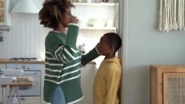 Smiling loving African American mother measuring height of child son at home, using metal ruler and pencil, marking top of head on wall, mom taking measurement while boy standing against flat surface